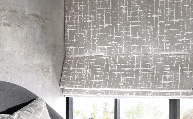 Textured grey bathroom Roman blind partially lowered over a large window with patterned fabric.