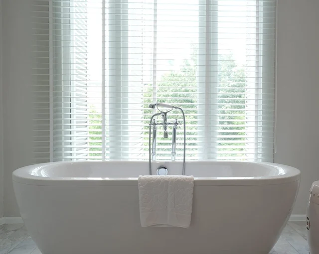 Freestanding bath in front of white wooden bathroom blinds filtering natural light