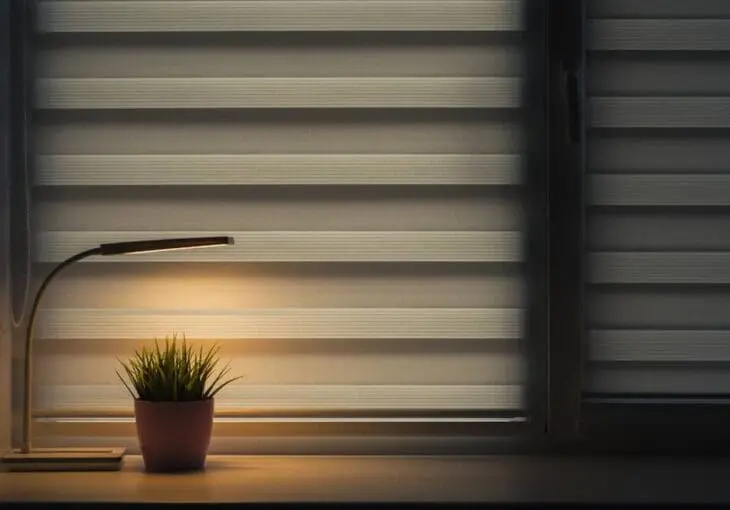 modern day and night blinds closed in a bedroom with soft lighting from a desk lamp and plant on the windowsill